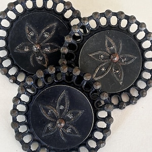 May include: Three circular black buttons featuring a central engraved five-petaled flower design. Each button has a dark metallic filigree border with small raised studs along the outer edge and a metallic center point.