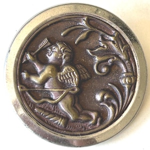 May include: A round, antique-style button with a detailed relief of Cupid aiming an arrow. The button features a bronze-colored Cupid with gold wings and a bow, set against a dark brown background, framed by a silver-colored metal edge.