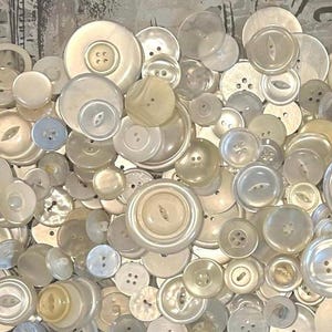 May include: A collection of various sizes and shapes of off-white and cream-colored buttons. The buttons have two or four holes, and some have a pearlescent sheen. The buttons are scattered across the image.
