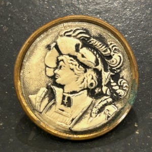 May include: A round, antique button with a detailed relief of a person in profile, wearing a hat and armor. The central image is off-white, set within a gold-colored metal frame. The button shows signs of age.