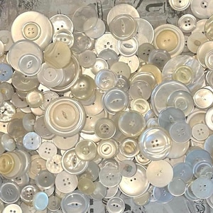 May include: A collection of various sizes and styles of off-white and pale-yellow buttons. The buttons are made of different materials, including mother-of-pearl, and have two or four holes. Some buttons have a raised center.