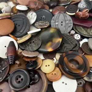 May include: A large collection of assorted buttons in various shades of brown, black, white, and gray. The buttons feature diverse textures, shapes, and sizes, including round, flat, and cylindrical designs with two or four holes, some with intricate patterns and smooth or matte finishes.