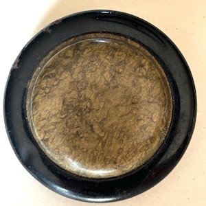May include: A round, antique-style compact with a black rim and a gold-toned center. The center has a textured, mottled appearance. The compact appears to be made of metal and has a glossy finish. The black rim shows some signs of wear.