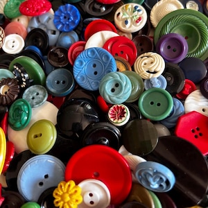 May include: A large collection of assorted sewing buttons in various shapes, sizes, and colours including red, blue, green, black, white, and yellow. The buttons feature diverse textures, patterns, and hole configurations, creating a dense, layered pile.