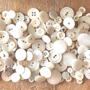 Peut inclure: Un assortiment de boutons blanc cassé de différentes formes, ronds et ovales, avec deux ou quatre trous. Les boutons sont disposés sur une surface en bois brun clair, présentant une variété de tailles et de textures.