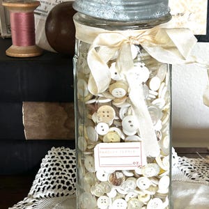 May include: A clear glass jar filled with various sizes and shades of off-white buttons. The jar has a metal lid and is tied with a cream-colored ribbon. A label reads "Parsley Flakes".