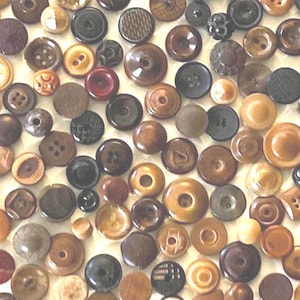 May include: A collection of assorted buttons in various shades of brown, tan, black, and cream. The buttons feature diverse textures, patterns, and hole configurations, including two-hole and four-hole designs, scattered across a flat surface.