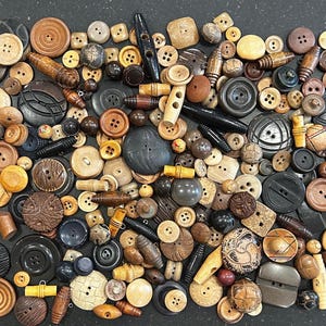 May include: A large collection of assorted buttons in various shapes, sizes, and materials, including wood, plastic, and metal. The buttons feature diverse textures, patterns, and colors such as brown, black, tan, and cream, scattered across a dark surface.