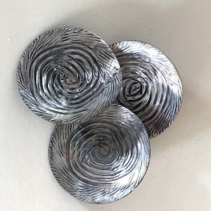 May include: Three circular buttons with a metallic silver finish. Each button features a distinct, raised swirling texture that radiates from the center toward the outer edge, creating a dynamic, light-reflecting pattern across the surface.