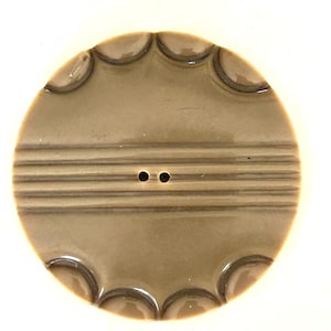 May include: A large, round, tan button with a scalloped edge and two small holes. The button has three horizontal, parallel lines across the center. The button is made of a smooth, shiny material.