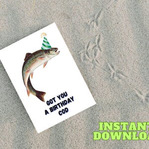 May include: A white birthday card featuring a fish wearing a party hat. The card has the text "GOT YOU A BIRTHDAY COD" in blue. The image is set on a sandy surface.