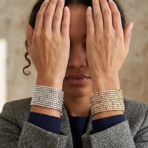 Puede incluir: Dos pulseras de múltiples hilos en las muñecas. Una pulsera está compuesta por cuentas metálicas de tono plateado dispuestas en varias filas, mientras que la otra tiene cuentas metálicas de tono dorado en un diseño similar.