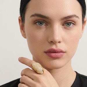 May include: A gold ring featuring a large, smooth, oval-shaped cream-colored stone set in a polished metal band. The stone has subtle, swirling light beige patterns across its surface, held securely by a curved, organic-shaped gold setting.