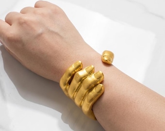 May include: A gold-toned cuff bracelet shaped like a human hand wrapping around the wrist. The design features four fingers resting across the top of the wrist and a separate thumb piece positioned on the opposite side of the wrist band.