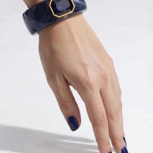 Può includere: Un bracciale rigido largo in resina marmorizzata blu scuro con una pietra centrale rettangolare sfaccettata blu scuro, incastonata in una cornice di metallo color oro. Il bracciale ha una finitura liscia e lucida e un design a fascia spessa.