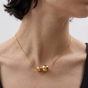 May include: A gold-toned necklace featuring a thin, smooth chain and two polished, spherical beads of different sizes positioned at the center. The larger sphere sits to the right of the smaller sphere, both reflecting light on their metallic surfaces.