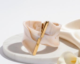 May include: A wide cuff bracelet featuring a marbled cream and beige resin body with a smooth, polished finish. The bracelet is accented by a vertical, rectangular gold-tone metal bar that serves as a central decorative element and hinge detail.