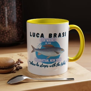 May include: White ceramic mug with a yellow interior and handle. The mug features the text "LUCA BRASI FISH MARKET" and a fish illustration. Additional text reads "Manhattan, New York" and "Where he sleeps with the fishes."