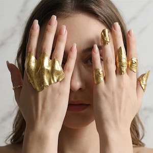 May include: A collection of gold-toned rings with irregular, textured surfaces. One large, wide ring covers multiple fingers, while several smaller rings are worn on individual fingers, including a nail-covering ring and various band styles.