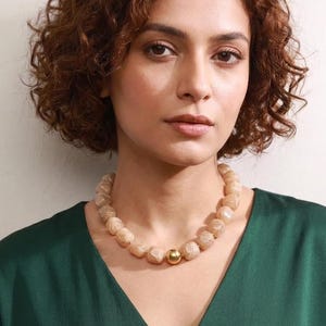 May include: A necklace featuring a series of light beige, marbled, cube-shaped beads strung together. The design is centered by a single, smooth, polished gold-tone spherical bead, creating a distinct contrast against the textured, square-shaped beads.