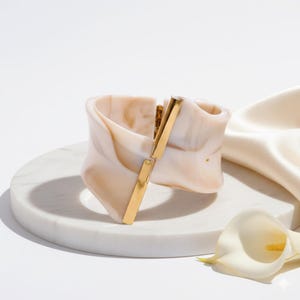 May include: A wide, marbled-effect cuff bracelet in shades of cream and tan, with a gold-tone clasp. The bracelet is displayed on a white marble surface, with a white flower and satin fabric in the background.