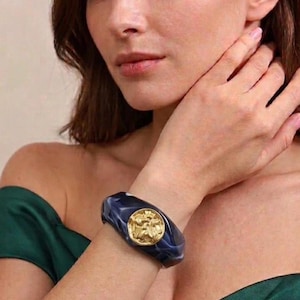 May include: A wide, dark blue and white marbled resin bangle bracelet featuring a circular, textured gold-tone metal emblem centred on the front. The bracelet has a smooth, polished finish and a thick, rounded profile that sits on the wrist.