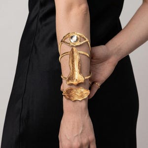 Op de afbeelding: Een goudkleurige armmanchetset bestaande uit drie afzonderlijke stukken: een oogvormige band met een heldere steen in het midden, een getextureerd neusvormig middendeel en een paar getextureerde lippen. Het metalen sieraad wordt op de onderarm gedragen en toont een gedetailleerd, sculpturaal ontwerp met een gepolijste afwerking die het licht vangt.