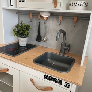 Könnte beinhalten: Eine weiße Spielzeugküche mit einer Holz-Arbeitsplatte, einem grauen Waschbecken und einer Mikrowelle. Sie enthält eine kleine Pflanze, eine Schüssel mit Buchstaben und Kochutensilien. Die Mikrowelle zeigt die Uhrzeit 12:30 an.