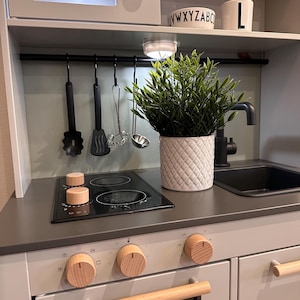Könnte beinhalten: Eine weiße Spielküche mit einem schwarzen Kochfeld, Backofen und Spüle. Das Kochfeld hat zwei Kochstellen mit Holzgriffen. Der Backofen hat einen Holzgriff und drei Holzgriffe. Die Spüle ist schwarz und hat einen Wasserhahn. Über dem Kochfeld befindet sich ein Regal mit einer weißen Tasse mit dem Buchstaben L darauf. Auf der Arbeitsplatte steht eine Pflanze in einem weißen Topf.