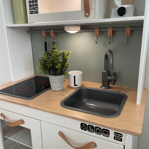 Könnte beinhalten: Weiße Spielküche mit einem grauen Spülbecken, einem schwarzen Kochfeld und einer Mikrowelle mit einem digitalen Display, das "12:30" anzeigt. Die Küche hat eine hellgrüne Rückwand und braune Ledergriffe. An der Rückwand befinden sich Haken zum Aufhängen von Utensilien.