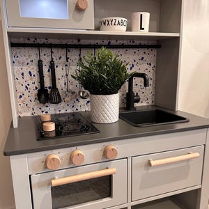 Könnte beinhalten: Eine graue Holzspielküche mit einer schwarzen Arbeitsplatte, einem schwarzen Spülbecken, einem weißen Backofen mit einem Holzgriff und einem schwarzen Kochfeld mit zwei Kochstellen. Die Küche hat ein Regal über dem Spülbecken mit einer weißen Tasse, auf der "L" steht, und einer weißen Schüssel, auf der "WXYZABC" steht.