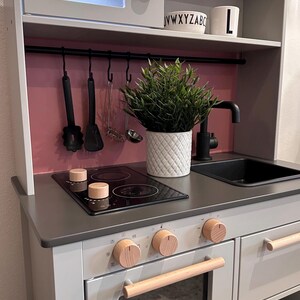 Könnte beinhalten: Graue und weiße Spielzeugküche aus Holz mit Mikrowelle, Herd, Backofen und Spüle. Das Set enthält Holzknöpfe, einen schwarzen Wasserhahn und einen weißen Topf mit einer grünen Pflanze. Küchenutensilien hängen über dem Herd.
