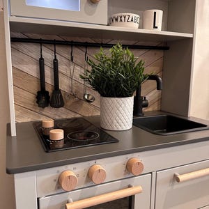Könnte beinhalten: Eine weiße Spielküche mit Mikrowelle, Herd, Backofen und Spüle. Die Küche hat eine Arbeitsplatte aus Holz und ein schwarzes Metallgestell zum Aufhängen von Utensilien. Auf der Arbeitsplatte steht eine kleine Pflanze in einem weißen Topf. Die Küche hat einen Holzgriff an der Backofentür und Holzknöpfe an Herd und Backofen. Die Küche hat ein Grau-Weiß-Farbschema.