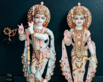 May include: Two white marble statues of deities, one playing a flute and the other with a raised hand. Both figures wear ornate gold crowns and are adorned with pastel-colored clothing and jewelry. The statues stand on square bases.