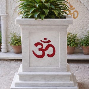 Puede incluir: Una jardinera de mármol blanco con un símbolo Om rojo, que contiene una planta verde. La jardinera es cuadrada y tiene una base escalonada. La planta tiene hojas largas y verdes con acentos rojos.