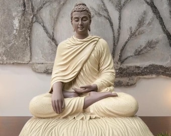 May include: A beige ceramic statue of Buddha sitting in meditation pose on a beige cushion. The statue is in a room with a white wall and a beige wall hanging behind it.