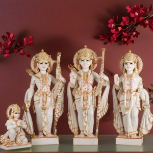 May include: White marble statues of Hindu deities, including Rama, Lakshmana, Sita, and Hanuman. The statues are intricately carved and feature gold accents. The statues are arranged in a row on a shelf.