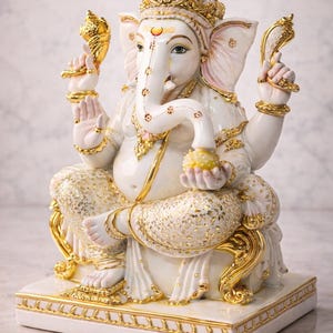 May include: A white and gold statue of the Hindu deity Ganesha, seated on a white base. The statue features intricate gold detailing on the crown, jewelry, and clothing. The elephant-headed figure holds a sweet treat in one hand.