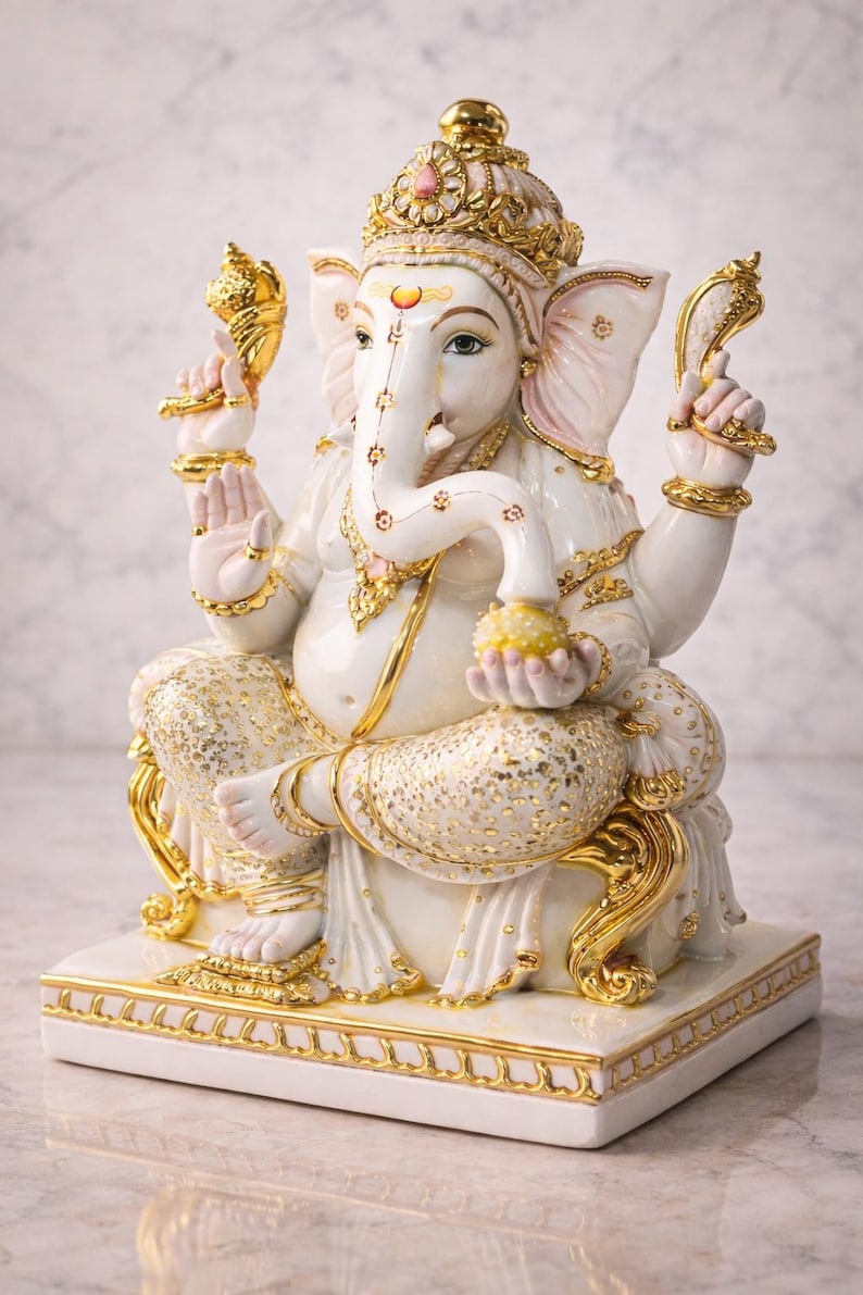 May include: A white and gold statue of the Hindu deity Ganesha, seated on a white base. The statue features intricate gold detailing on the crown, jewelry, and clothing. The elephant-headed figure holds a sweet treat in one hand.