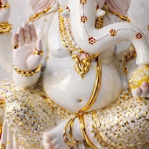 May include: A white ceramic statue of the deity Ganesha featuring gold-painted accents on the jewelry, clothing, and trunk. The figure is seated in a cross-legged position, holding a yellow orb, with intricate floral patterns on the trunk.