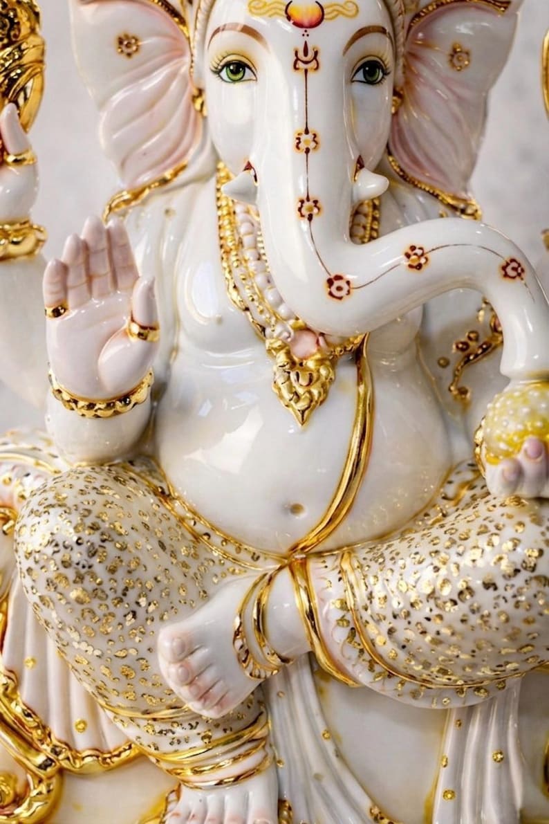 May include: A white ceramic statue of the deity Ganesha featuring gold-painted accents on the jewelry, clothing, and trunk. The figure is seated in a cross-legged position, holding a yellow orb, with intricate floral patterns on the trunk.