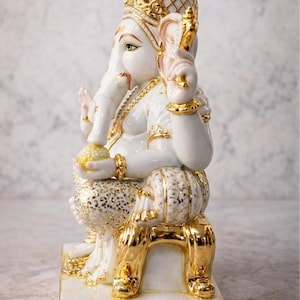 May include: A white and gold ceramic statue of the Hindu deity Ganesha. The statue features intricate gold detailing on the crown, jewelry, and clothing. Ganesha is seated on a gold chair, holding a yellow orb.