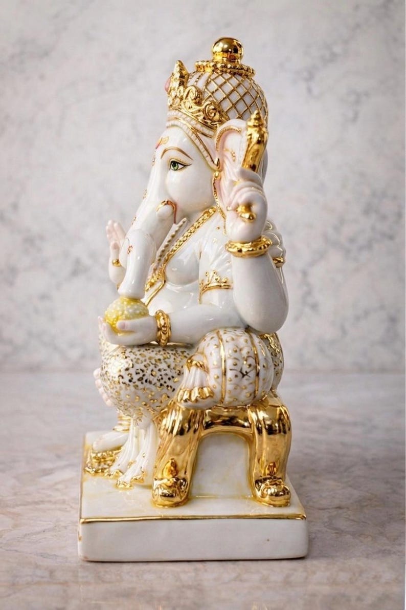 May include: A white and gold ceramic statue of the Hindu deity Ganesha. The statue features intricate gold detailing on the crown, jewelry, and clothing. Ganesha is seated on a gold chair, holding a yellow orb.