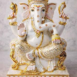 May include: A white ceramic statue of Ganesha seated on a base, featuring gold-painted accents on the crown, jewelry, and clothing. The figure has four arms, holding a conch shell and a lotus, with one hand raised in a gesture of blessing.