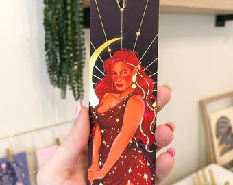 May include: A bookmark featuring a woman with red hair and a gold crown, wearing a red dress with gold stars. The background is black with gold stars and a crescent moon.