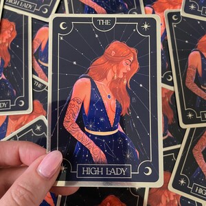 May include: A tarot card with a gold border and a dark blue background. The card depicts a woman with red hair wearing a blue dress with stars and constellations. The card is titled "The High Lady".