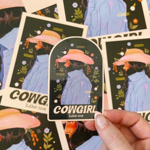 May include: A sticker with a black background and a woman wearing a pink cowboy hat and a blue denim jacket. The sticker has the text "COWGIRL Like me" in white letters.