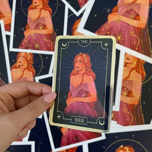 Op de afbeelding: Een gouden tarotkaart met een zwarte achtergrond en een vrouw in een paarse jurk met gouden sterren. De kaart heeft de tekst "THE SEER" onderaan.