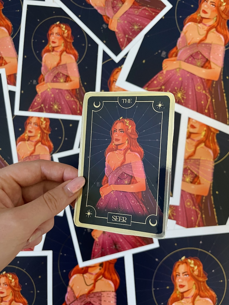 Op de afbeelding: Een gouden tarotkaart met een zwarte achtergrond en een vrouw in een paarse jurk met gouden sterren. De kaart heeft de tekst "THE SEER" onderaan.