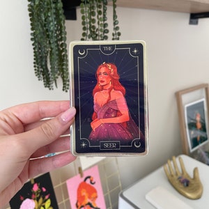 Op de afbeelding: Een tarotkaart met een gouden rand en een blauwe achtergrond. De kaart toont een vrouw met rood haar die een paarse jurk met gouden sterren draagt. De kaart heeft onderaan de tekst "THE SEER".